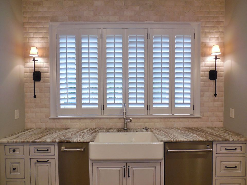 Kitchen with white plantation shutters over a sink, surrounded by beige stone backsplash and cream cabinets.