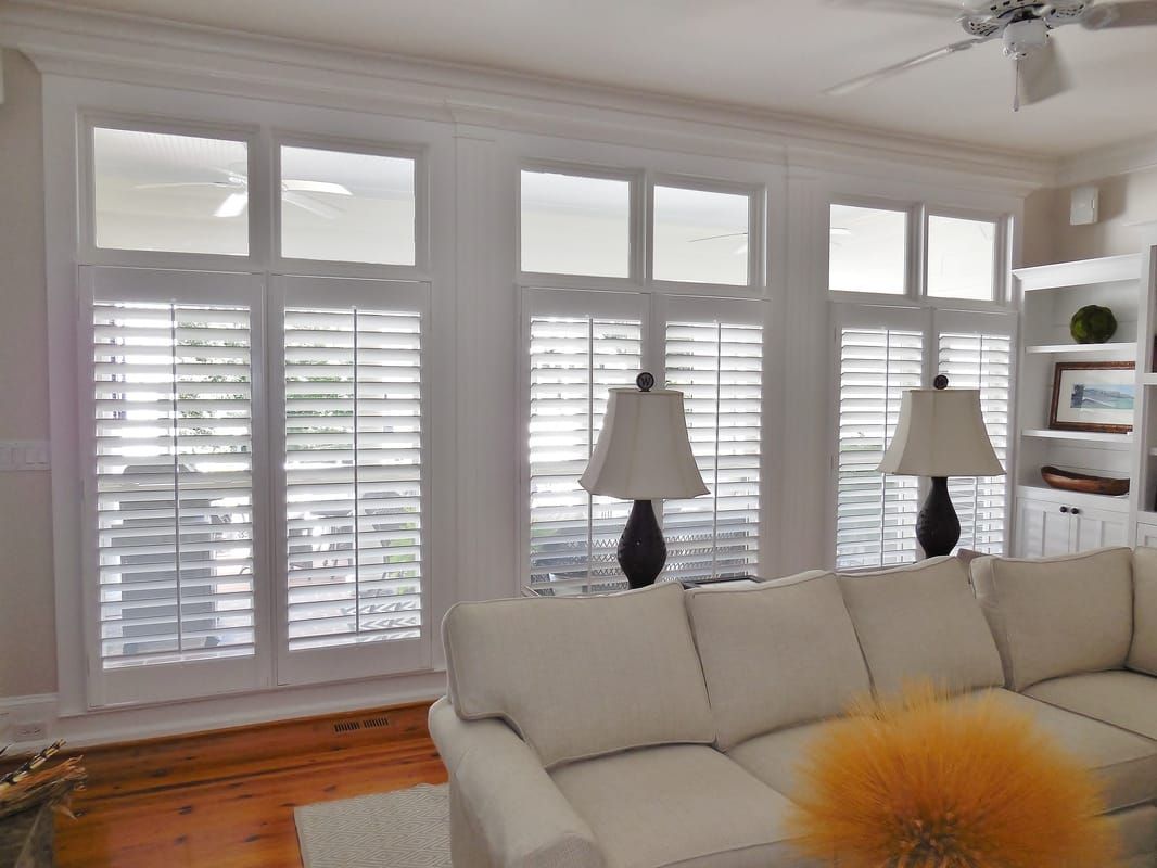 Bright living room with white shutters, beige sofa, wood floors, and lamps by the windows.