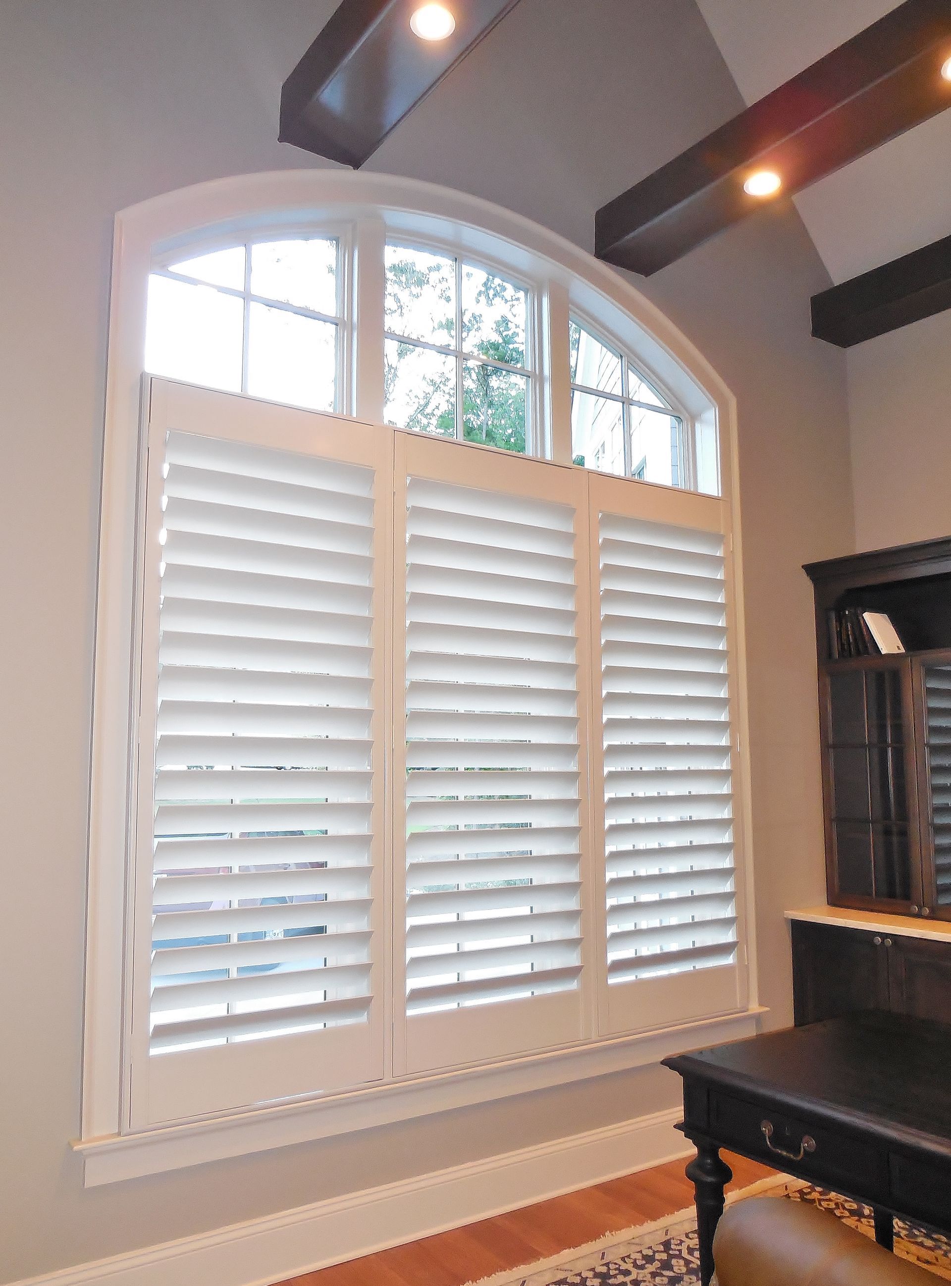 White plantation shutters beneath an arched window in a room with dark ceiling beams and a black piano