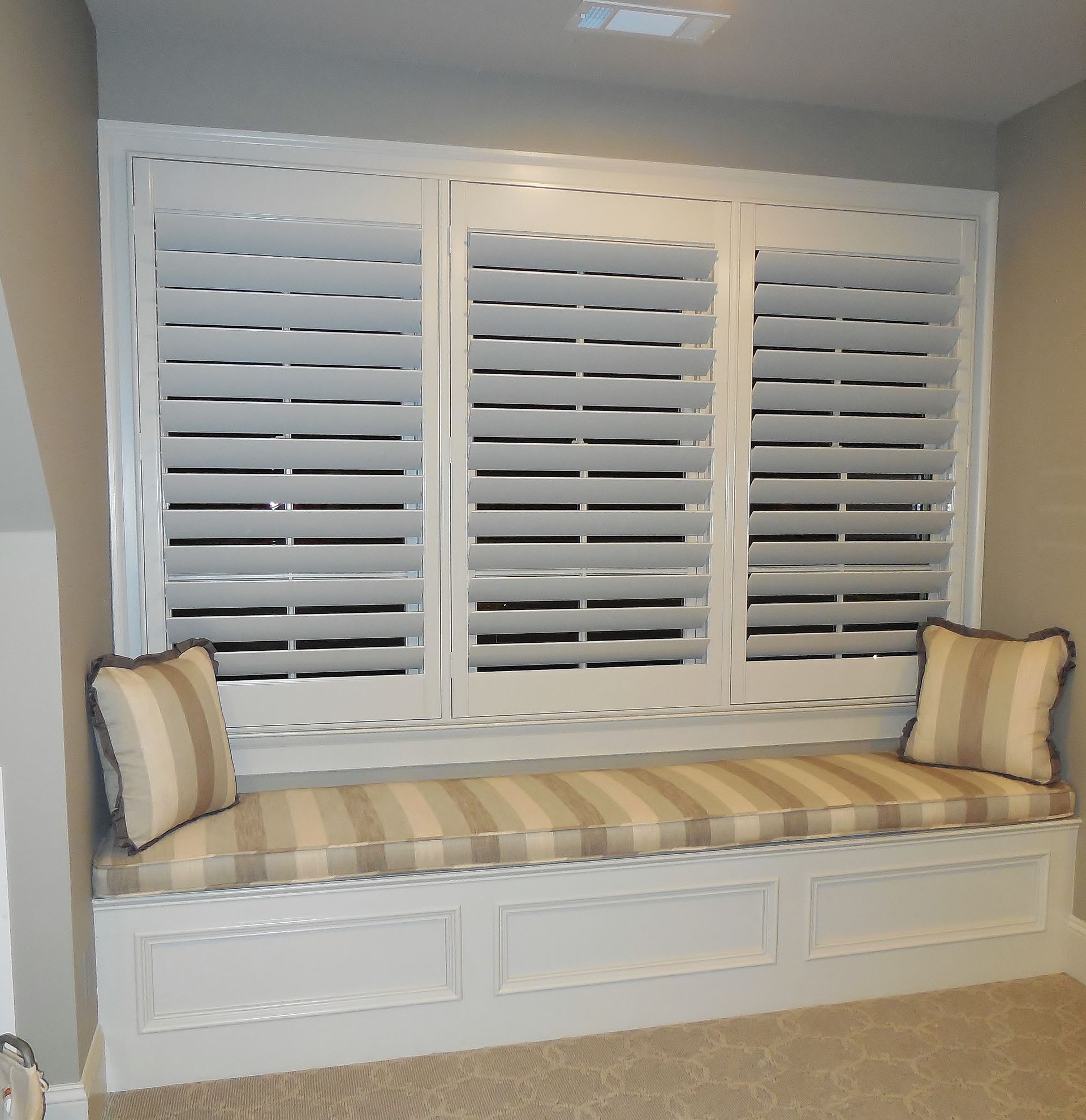 White built-in window seat with striped cushion and pillows beneath shuttered windows in a light room