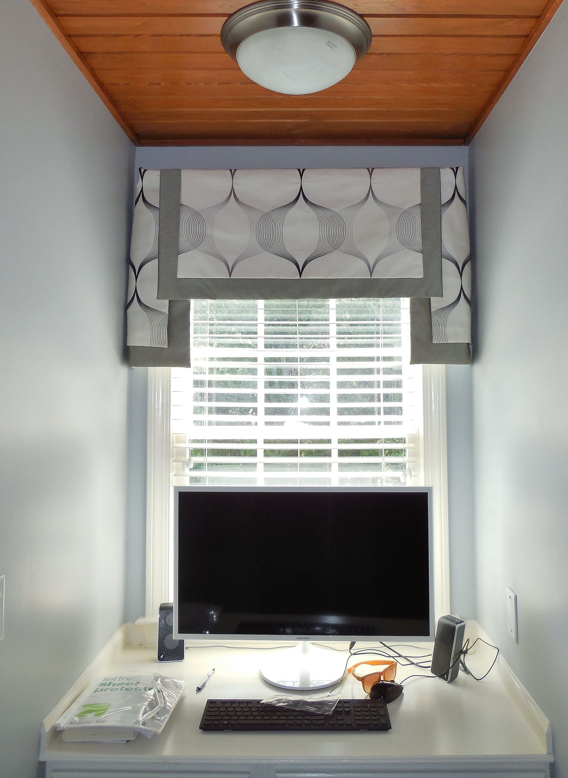 Compact desk with a laptop beneath a window with blinds and decorative valance in a narrow room.