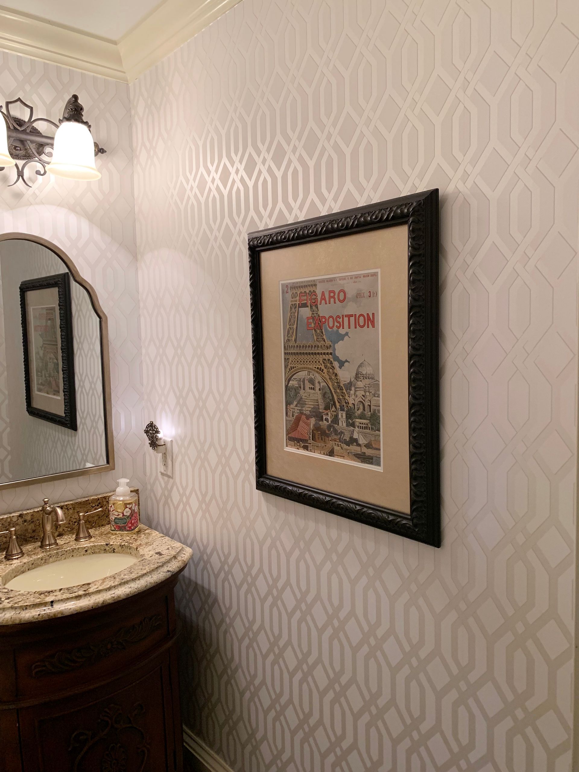 Small bathroom corner with ornate vanity, mirror, wall sconce, and framed art on patterned wallpaper.