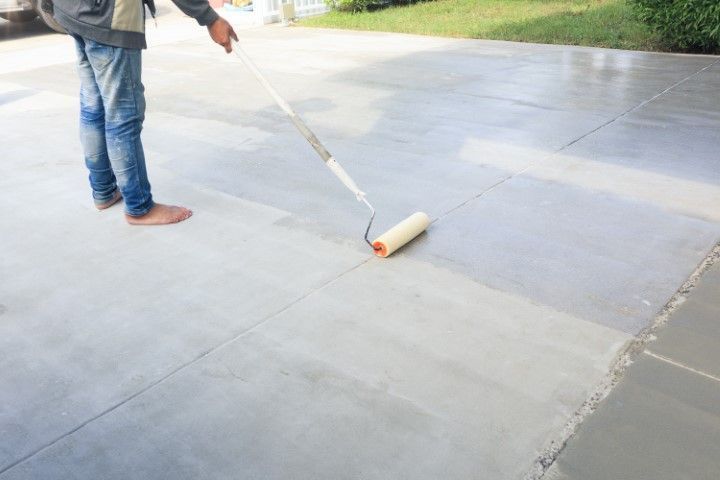 Worker applying a protective sealant to a grey concrete driveway.