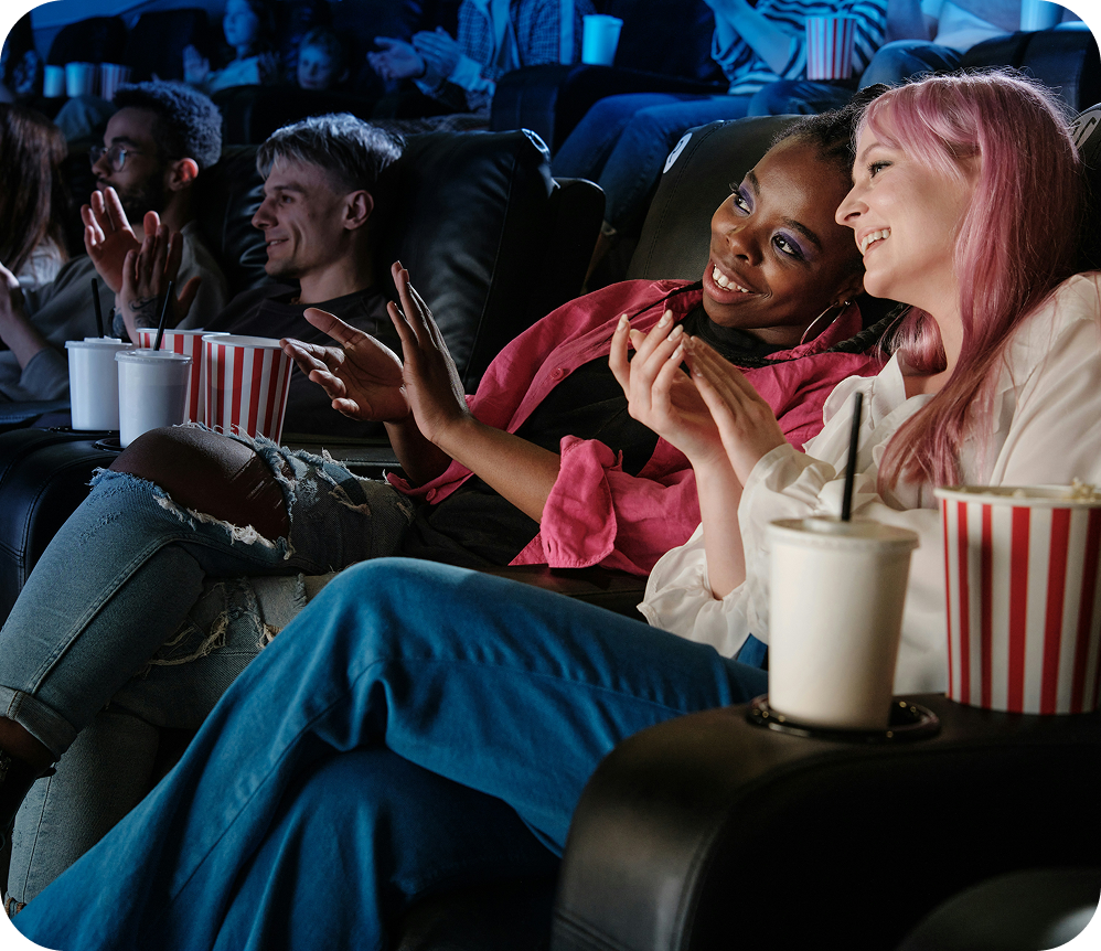 Friends chatting and laughing in a dim movie theater with popcorn and drinks during a film.