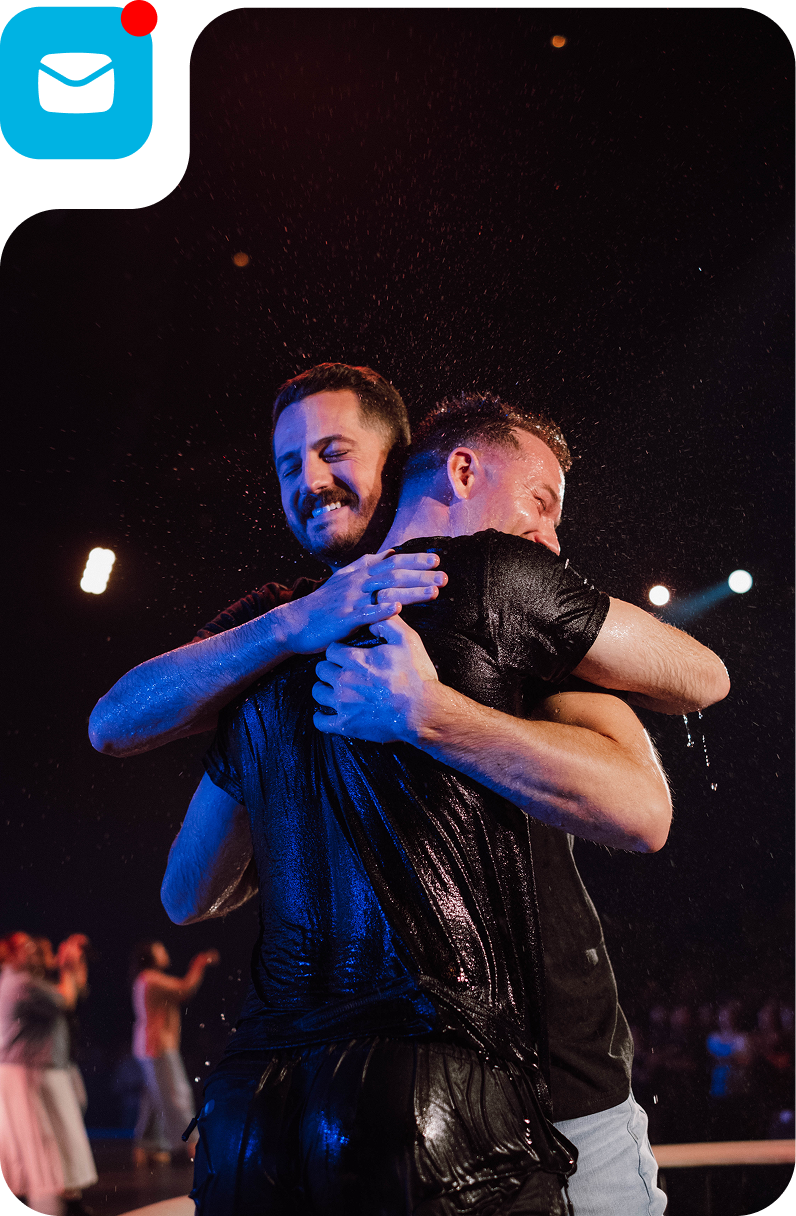 Two people hugging onstage under blue and warm lights, with a crowd in the background