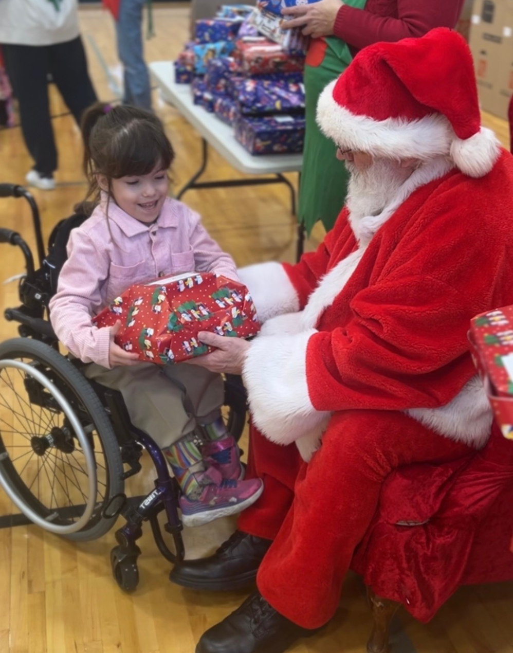 Child in wheelchair receives a wrapped gift from Santa in a festive indoor setting.