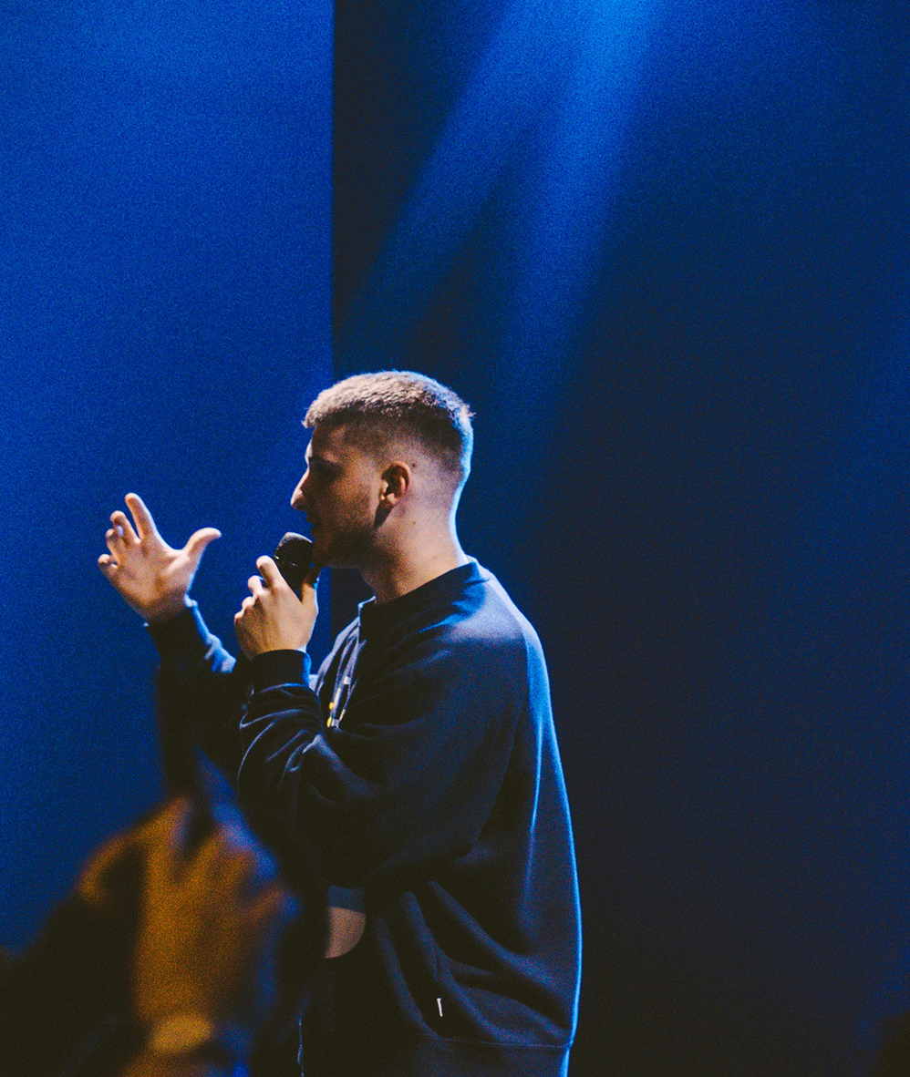 Singer holding a microphone on a blue-lit stage, gesturing with one hand