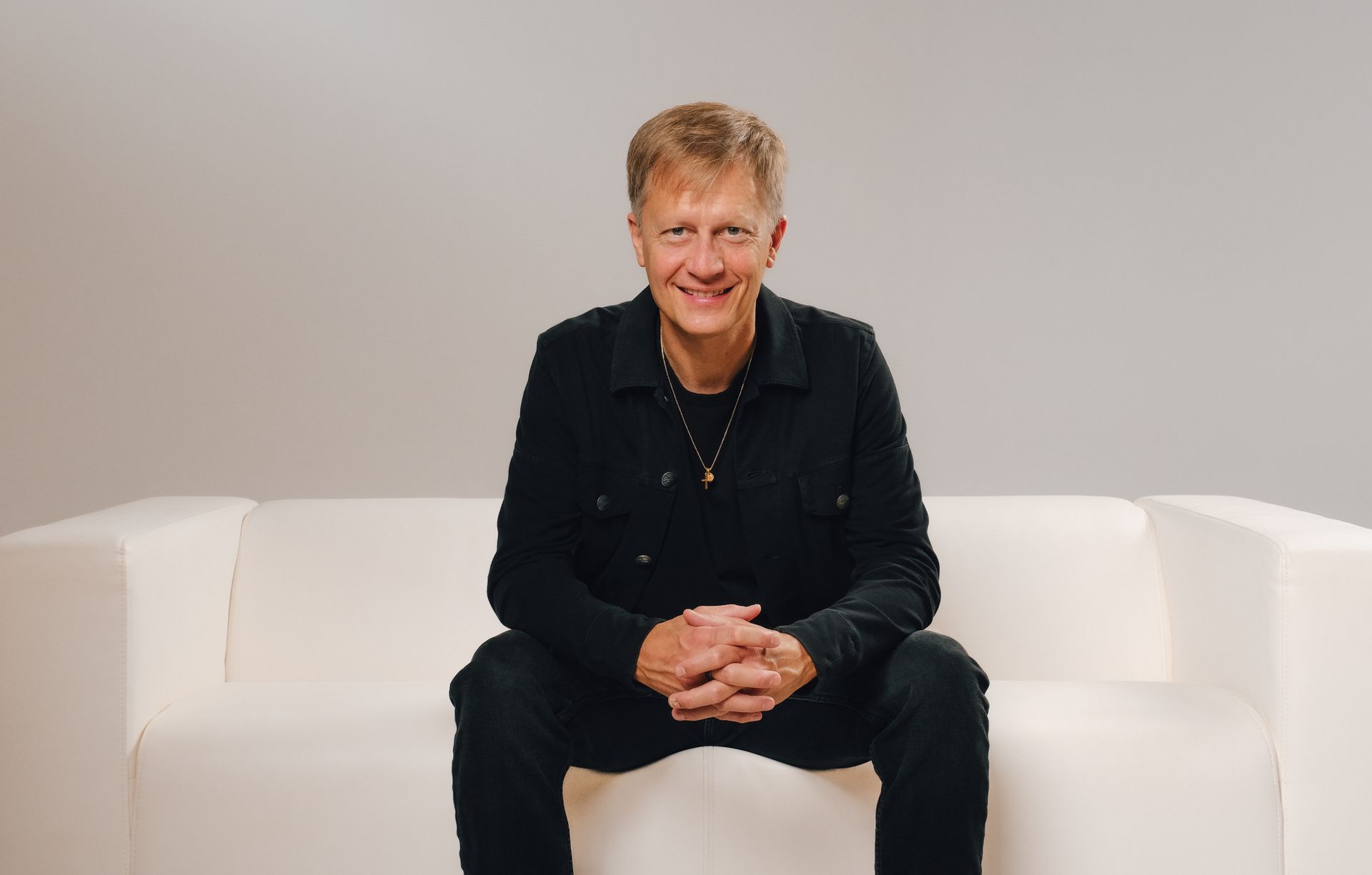 Smiling man in a black leather jacket, seated indoors against a softly blurred background