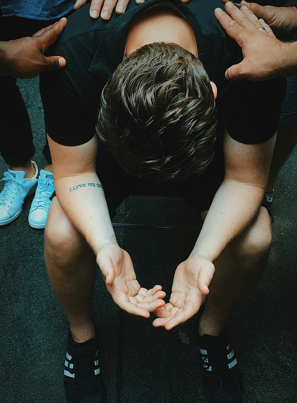 Exhausted runner sitting on pavement with head down, hands clasped, surrounded by teammates' hands