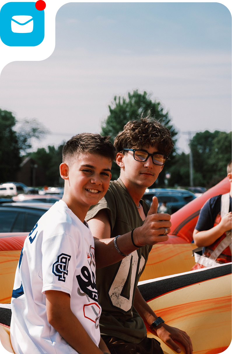 Two young men smiling beside a red car, one giving a thumbs-up outdoors.