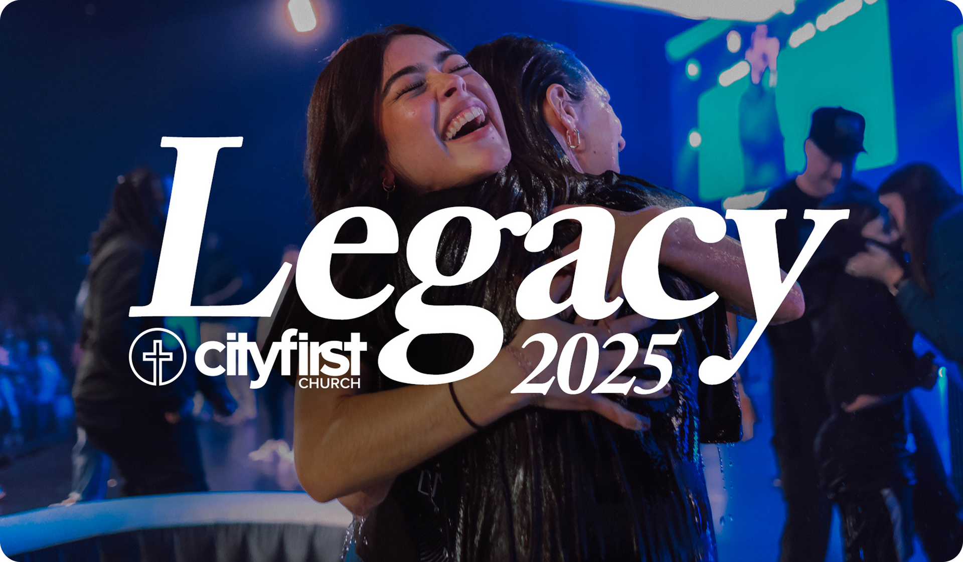 Two women hugging at a church event with “Legacy 2025” and CityFirst branding over a blue-lit crowd