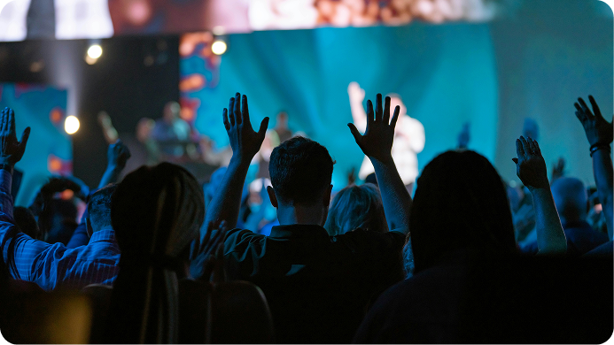 Silhouetted audience raising hands at a concert, with blue stage lights and blurred performers in the background