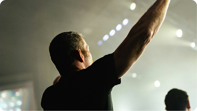 Person seen from behind raising an arm in a dim concert venue with bright stage lights.