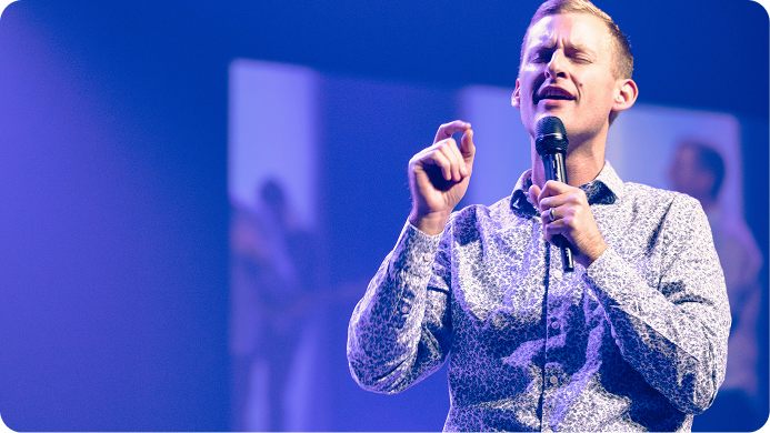 Speaker on stage holding a microphone, gesturing under blue and purple concert lighting.