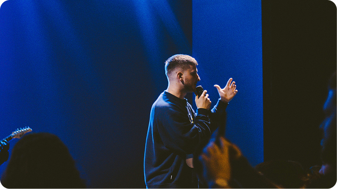 Man speaking onstage into a microphone, with blue lighting and blurred audience silhouettes in front