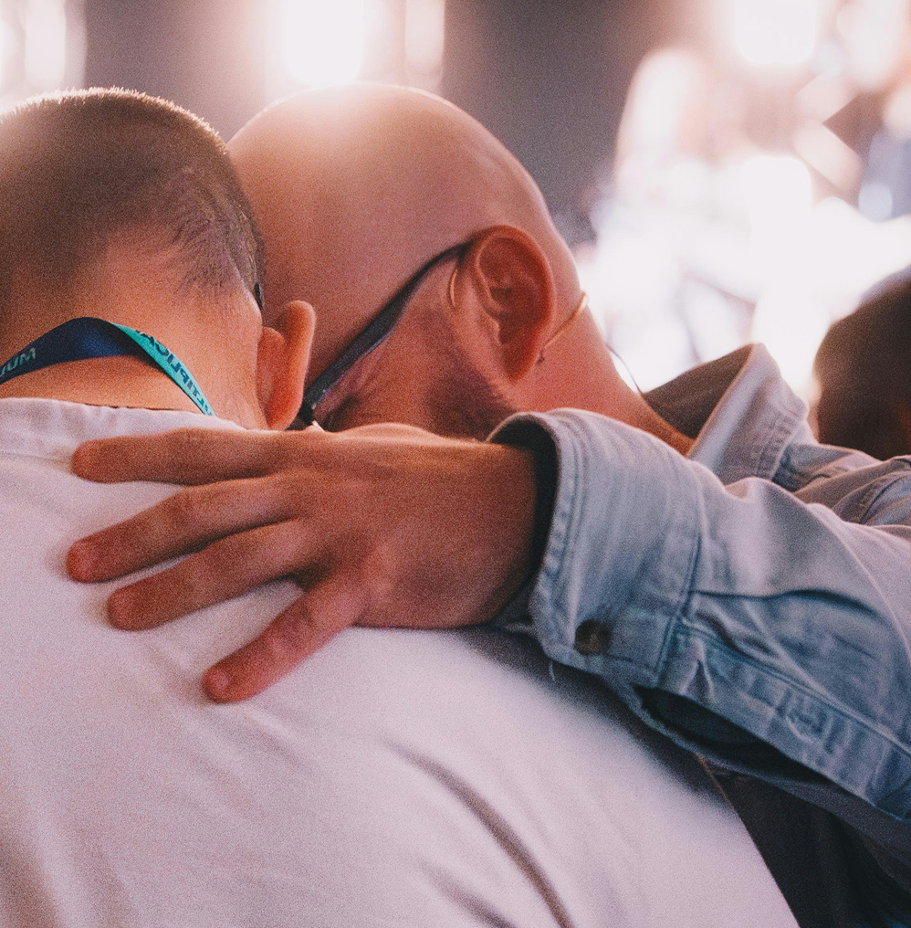 Two people embracing closely, one hand resting on the other's shoulder in warm light