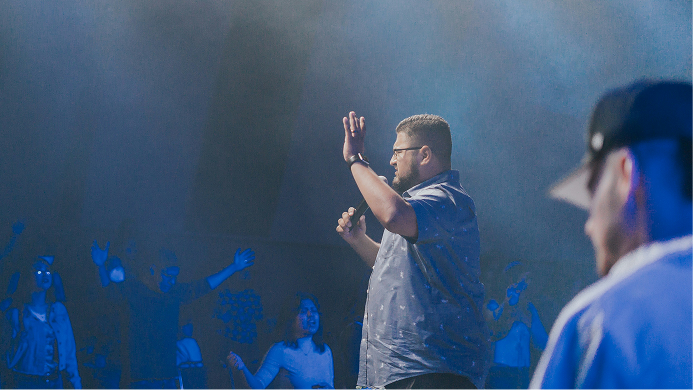 Man speaking on stage into a microphone, raising a hand amid blue stage lighting and audience silhouettes.
