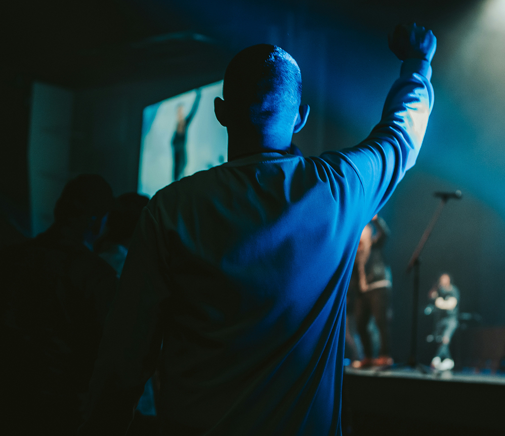 Person with raised arm in a blue-lit room, facing a stage or screen in a dark venue.
