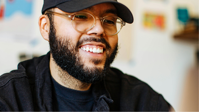 Smiling man in a black cap and glasses, wearing a dark jacket in a bright indoor setting