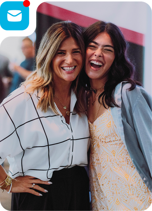 Two smiling women standing close together outdoors, one in a white grid shirt and one in a patterned top.