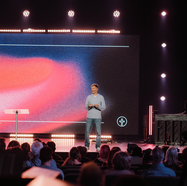 Speaker on stage in front of a large screen, addressing an audience under pink and purple lights