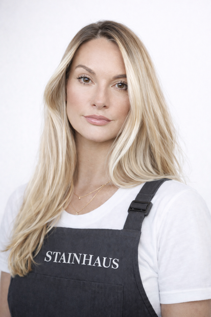 Blonde woman in a white T-shirt and dark apron with “STAINHAUS” text, standing against a light background