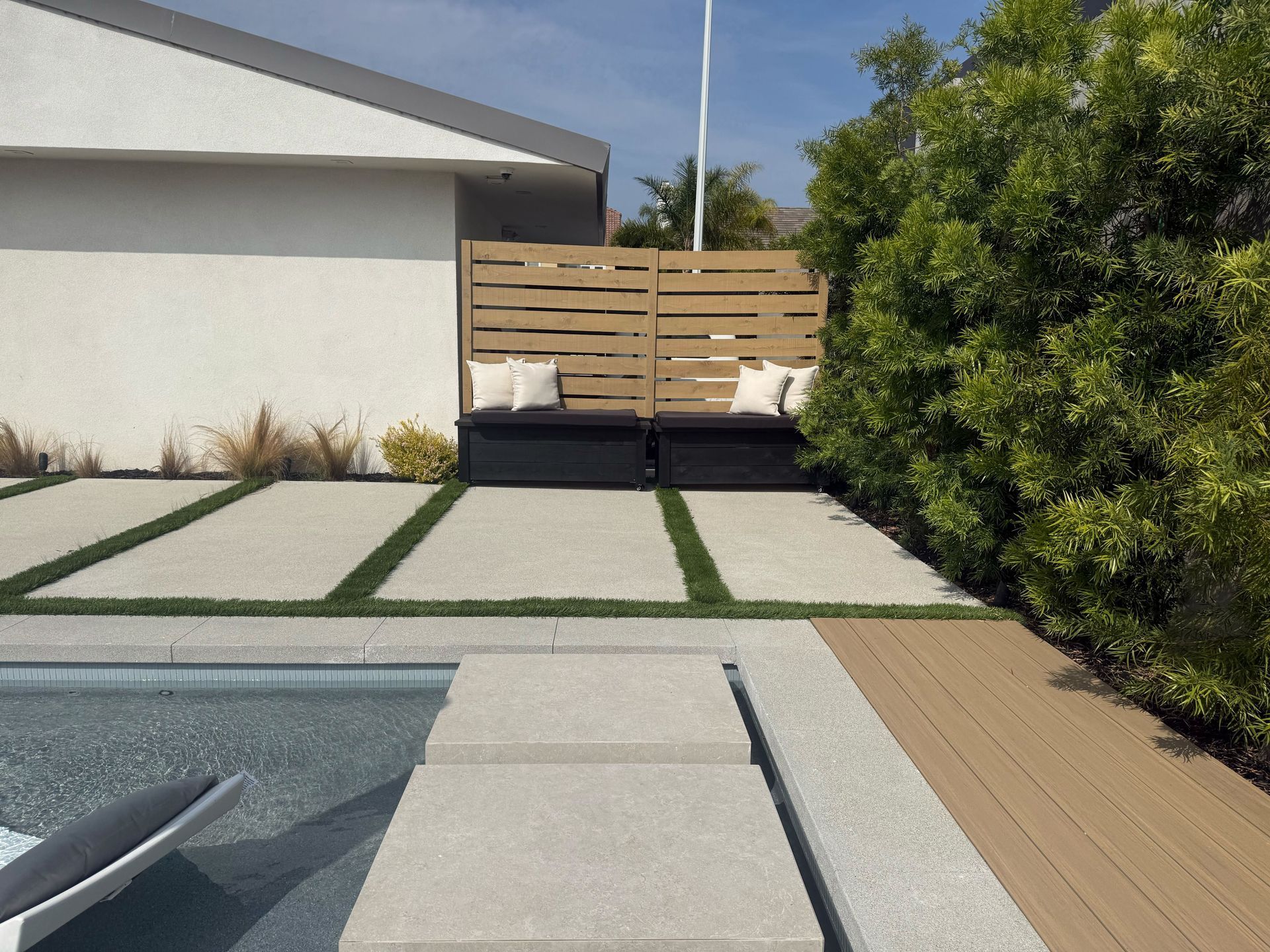 Backyard pool deck with gray pavers, wooden fence, and green shrubs beside a house