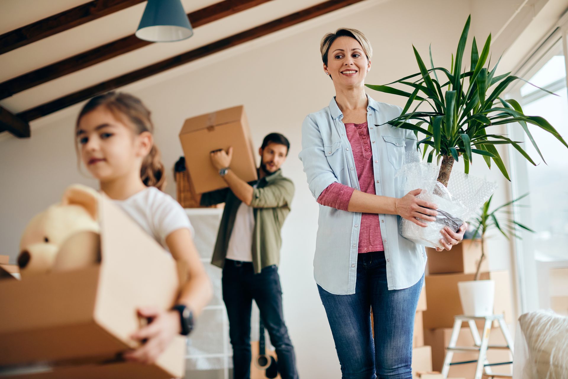 Family carrying boxes in a bright room, with a woman holding a potted plant during a move
