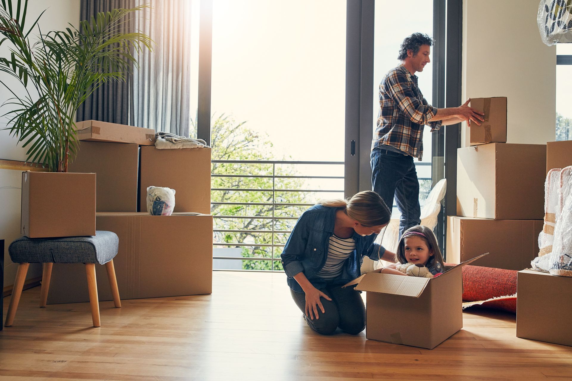 Family unpacking moving boxes in a bright living room; one adult kneels with a child while another stacks boxes.