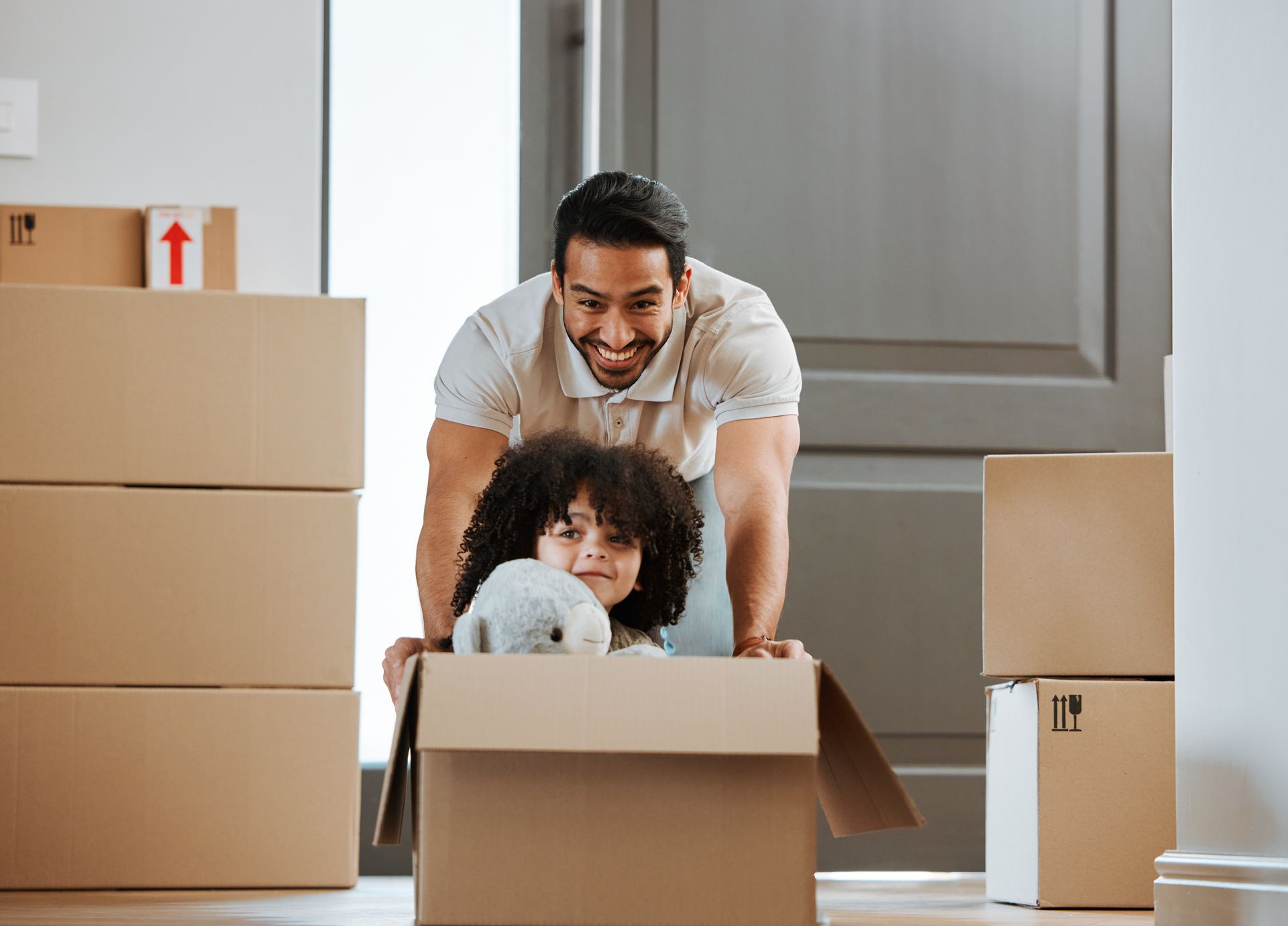 Man and child smiling in moving boxes, with child holding a stuffed toy in a bright room