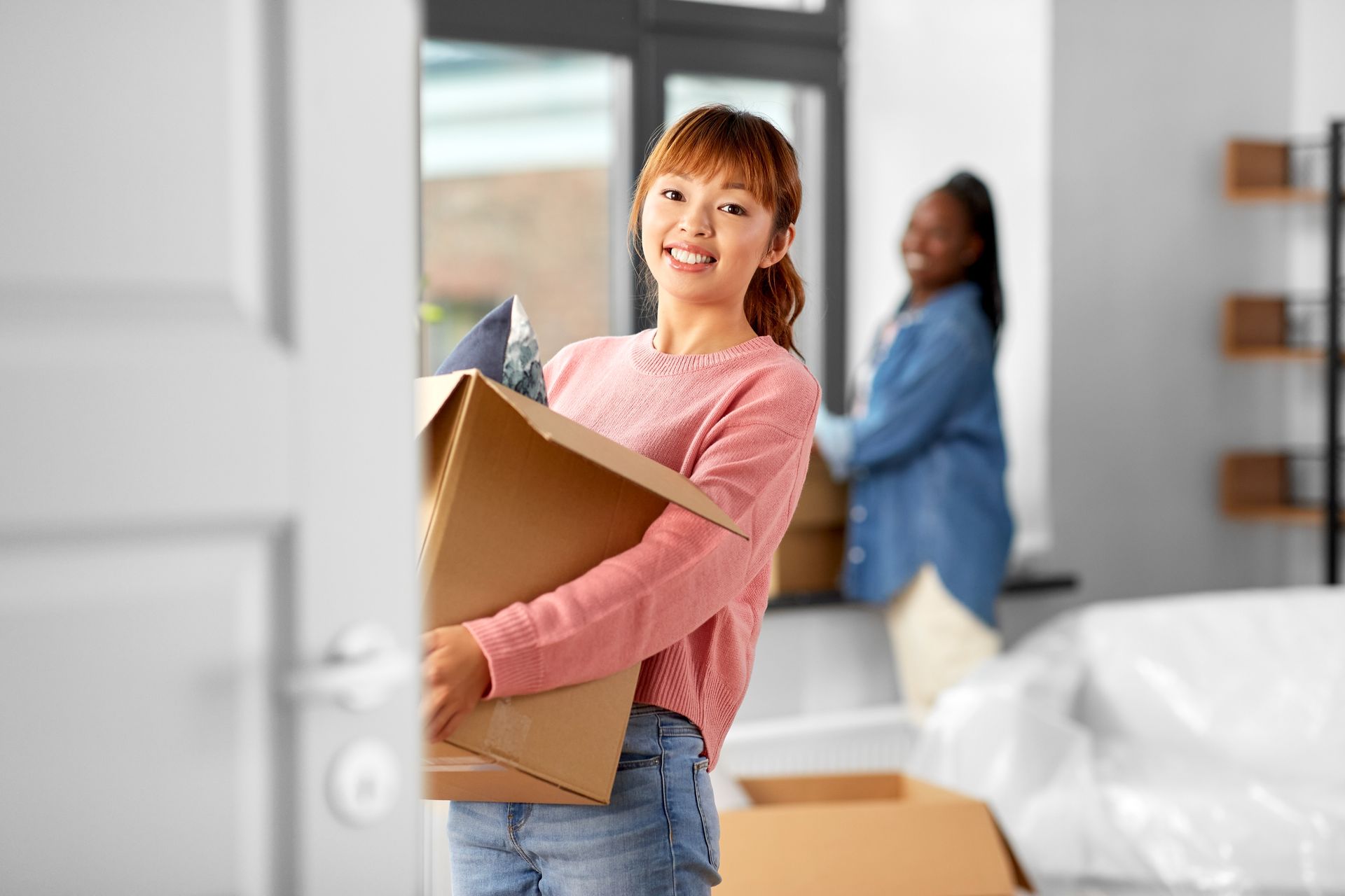 Person carrying a cardboard box into a bright room while another stands in the background.