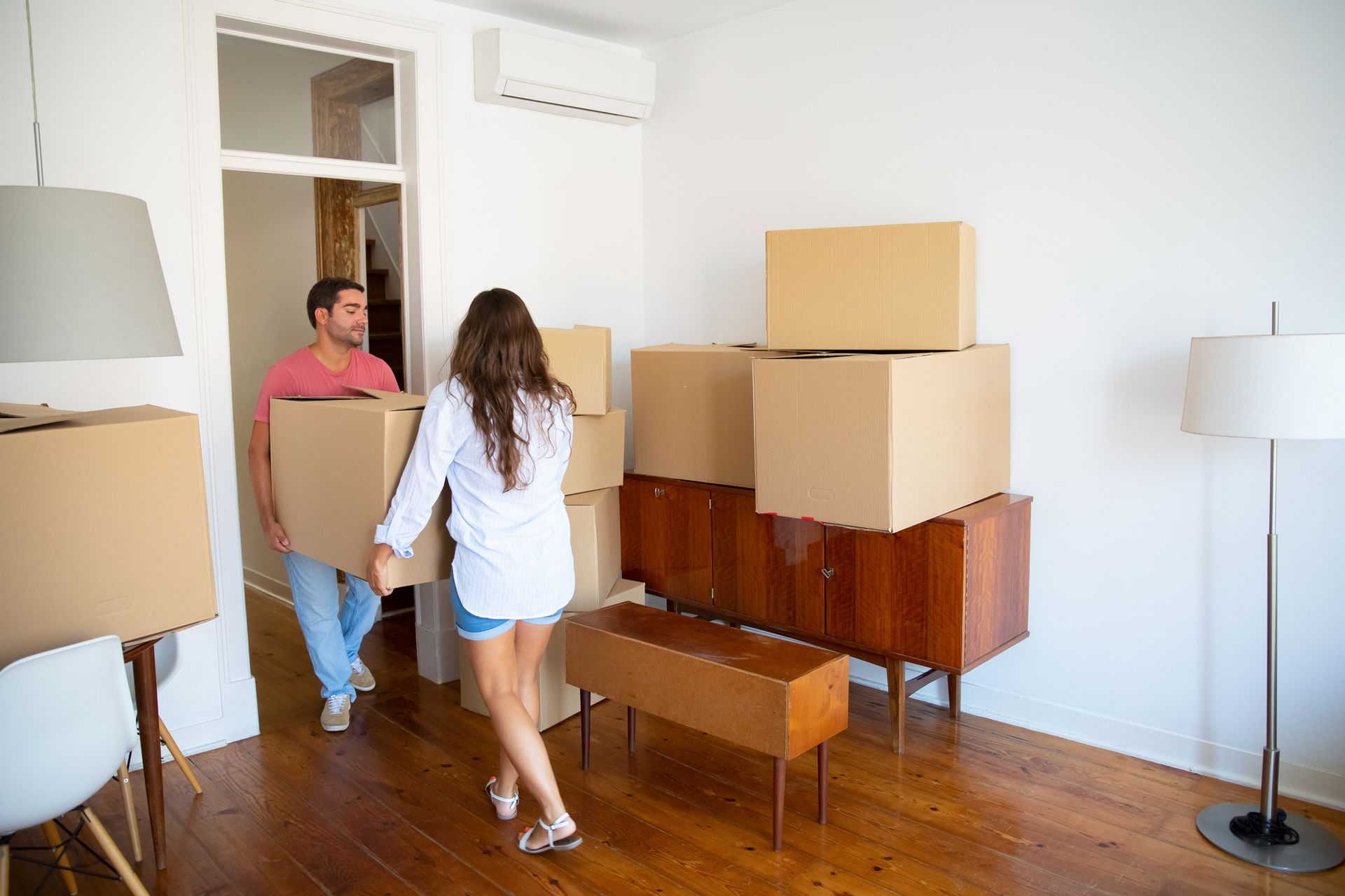 Two people move cardboard boxes in a bright room with hardwood floors and a floor lamp.