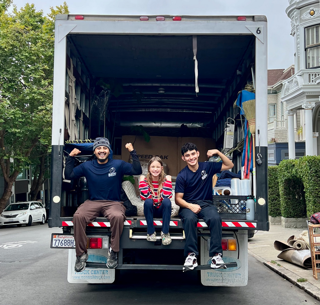 Three people sit on the open back of a box truck parked on a city street.