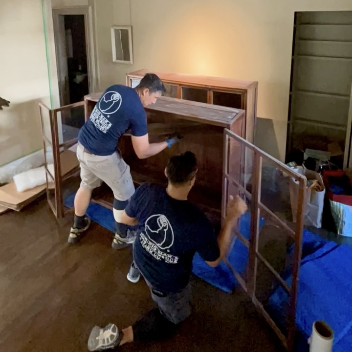 Two movers dismantling a wooden cabinet in a room. One stands, the other kneels.