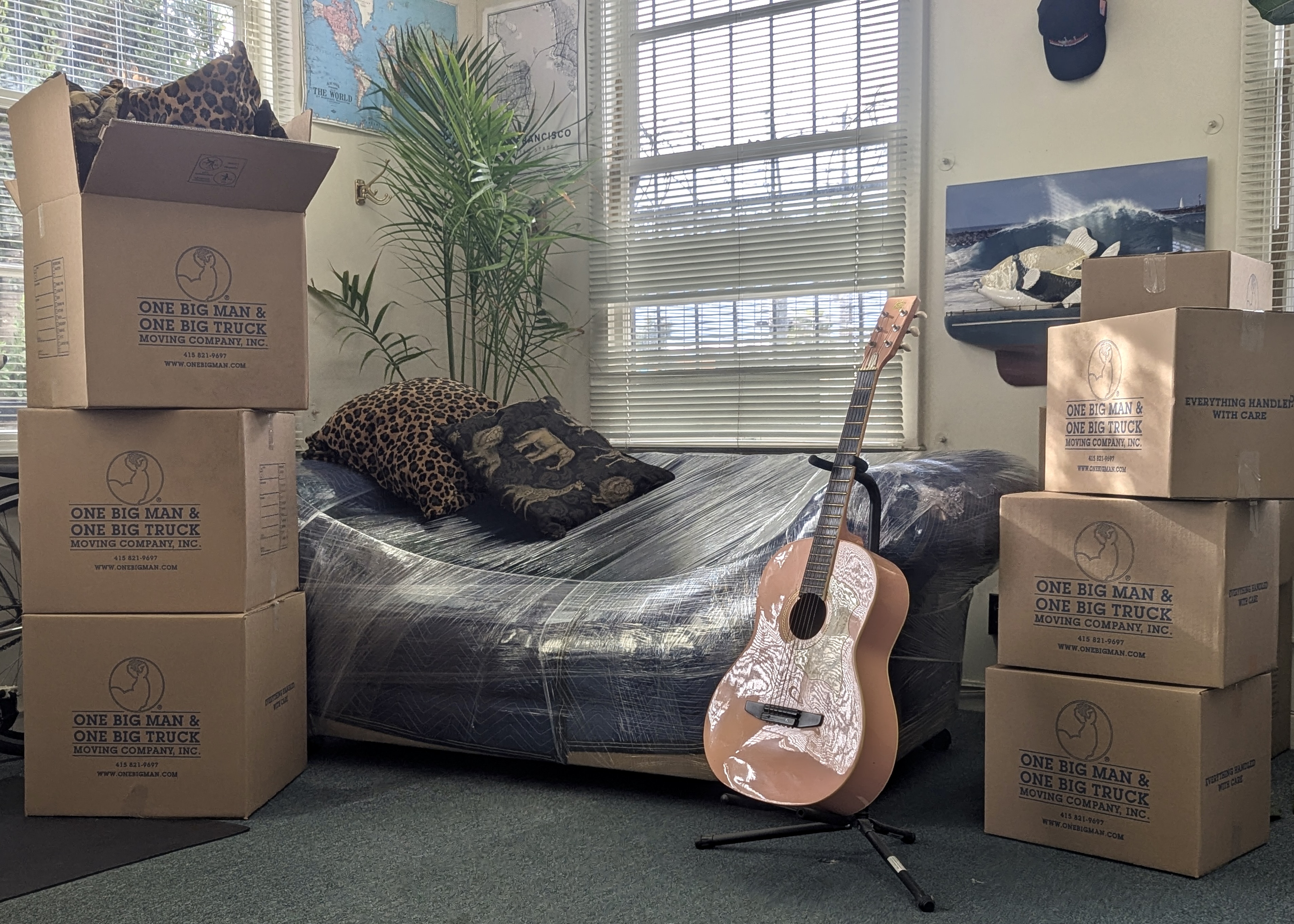 Boxes stacked next to a guitar on a stand and a bean bag chair in a room with a window.