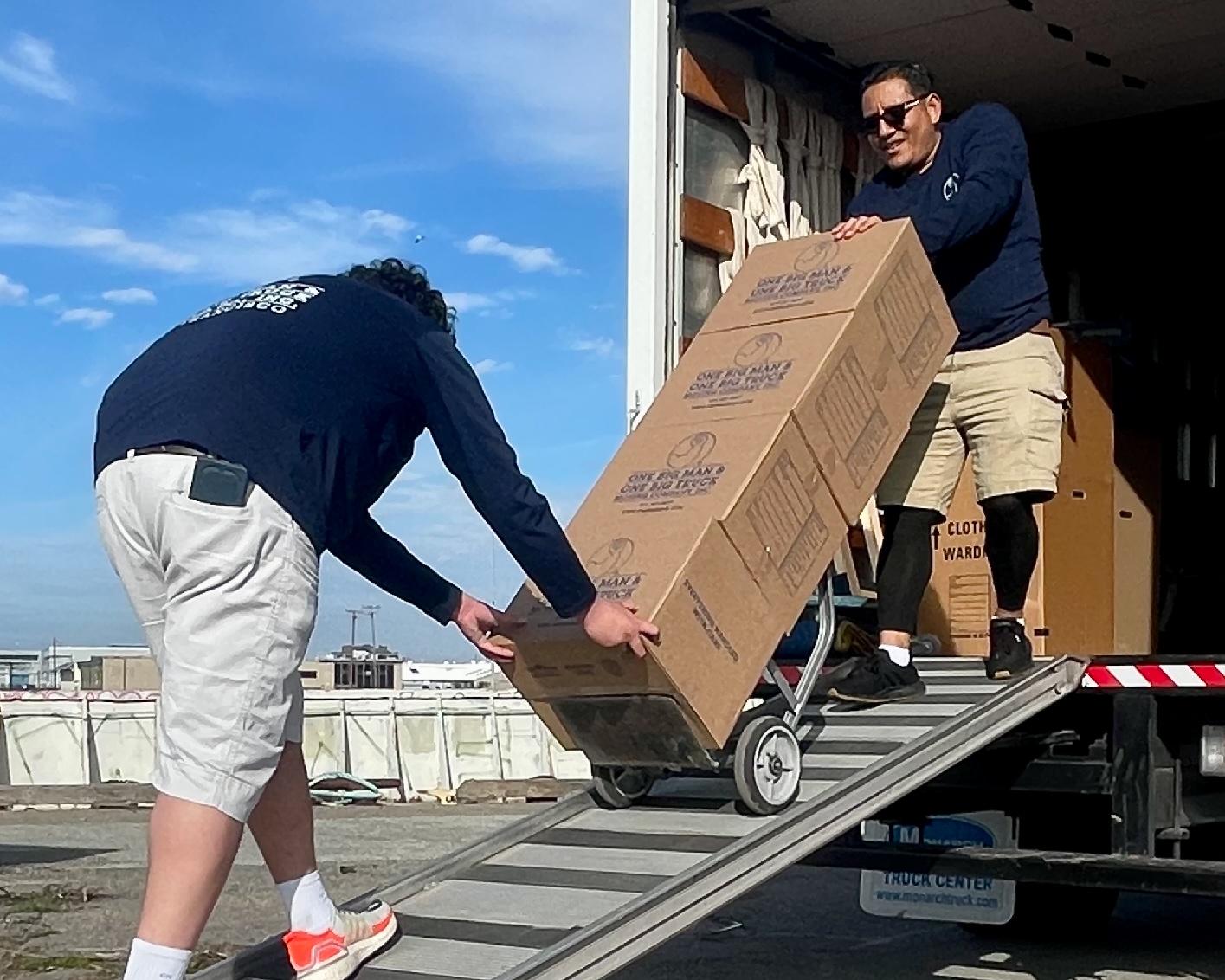 Two people loading large boxes from a truck onto a ramp. They wear shorts and are working together.