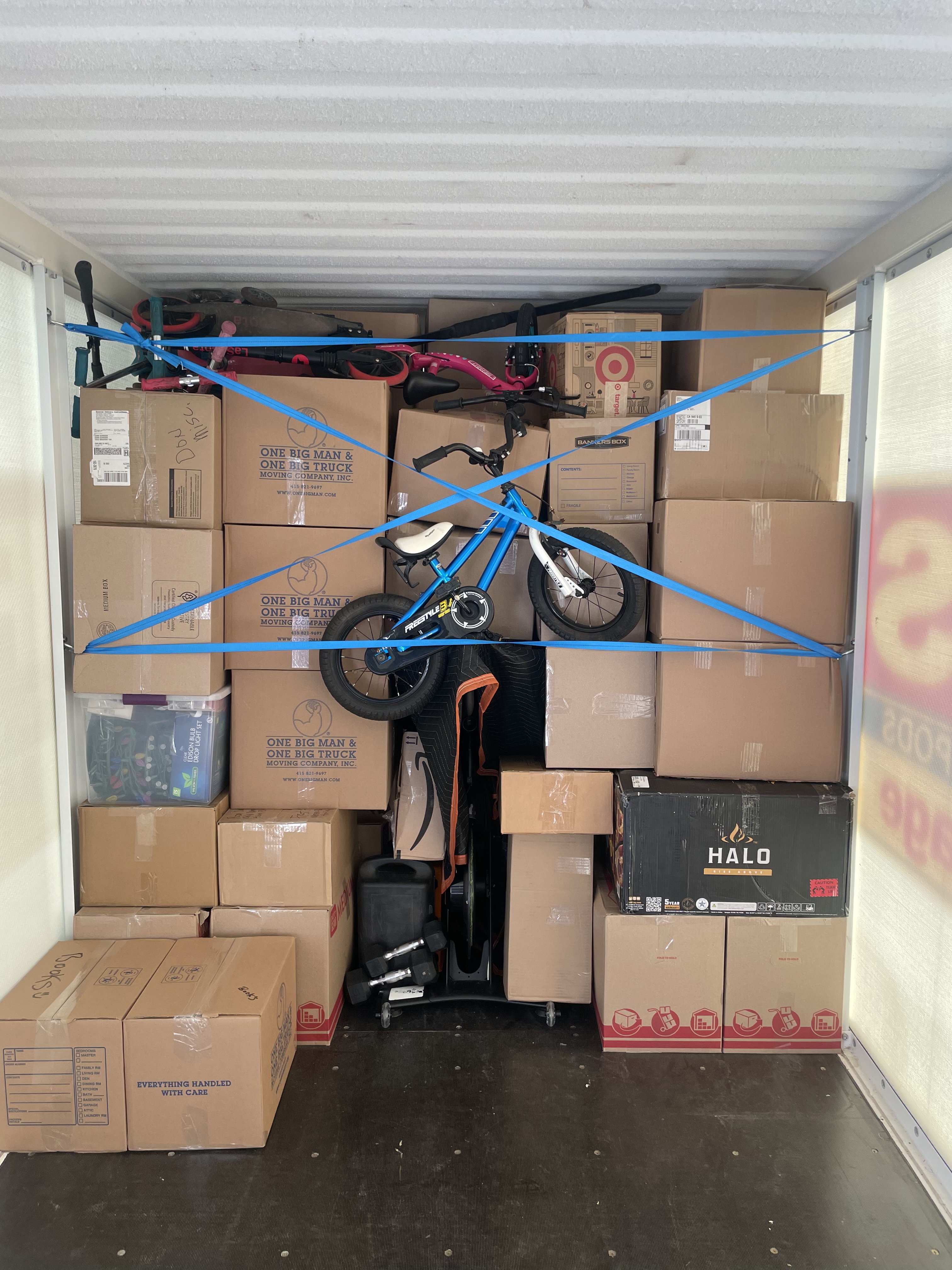 A view inside a storage unit packed with cardboard boxes, a blue bicycle, and other items secured by blue straps.