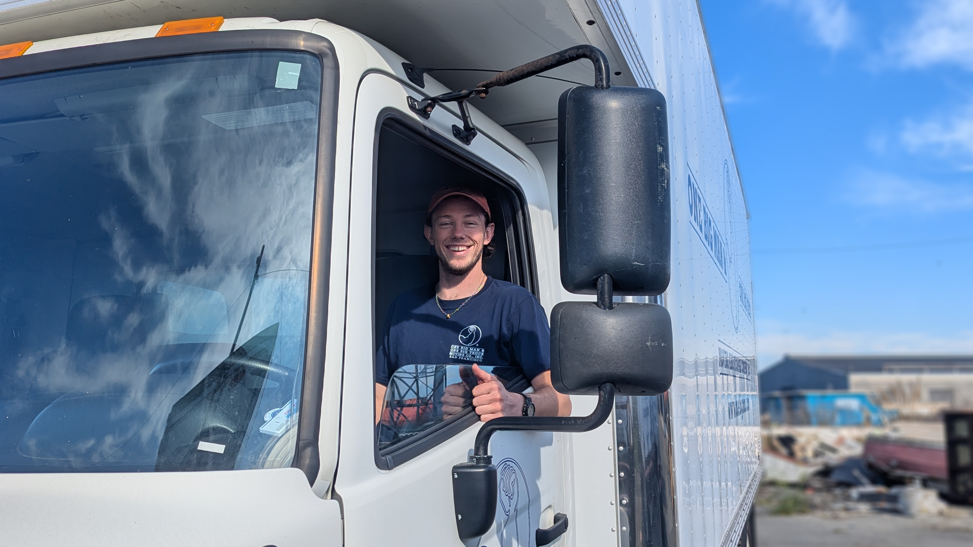 Person smiling and giving a thumbs up from inside a truck cab. Blue sky in background.
