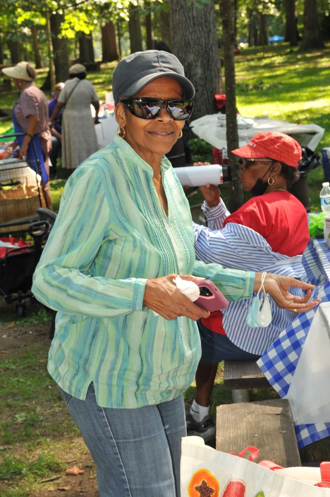 Woman in sunglasses holds a cup at an outdoor park gathering with tables and people nearby