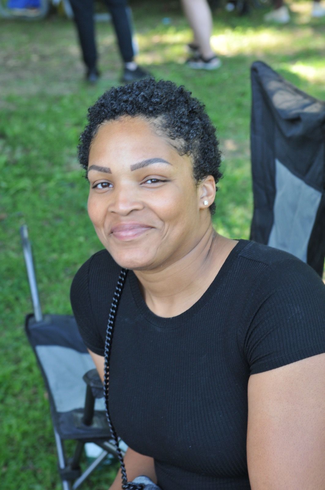 Smiling woman seated outdoors in a black top on a grassy lawn, with folding chairs in the background