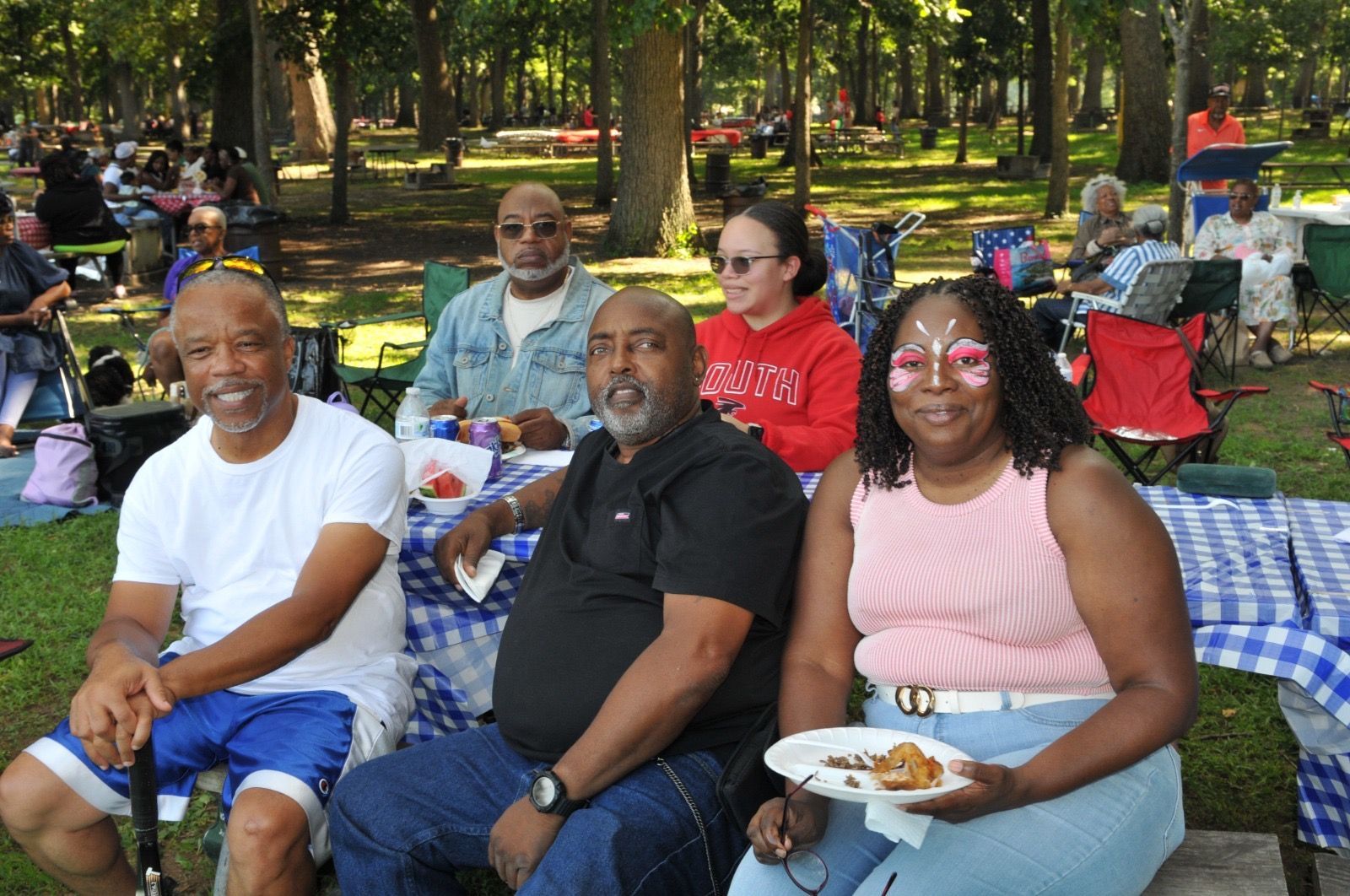 People seated on blankets at a park picnic, eating and relaxing under the trees.