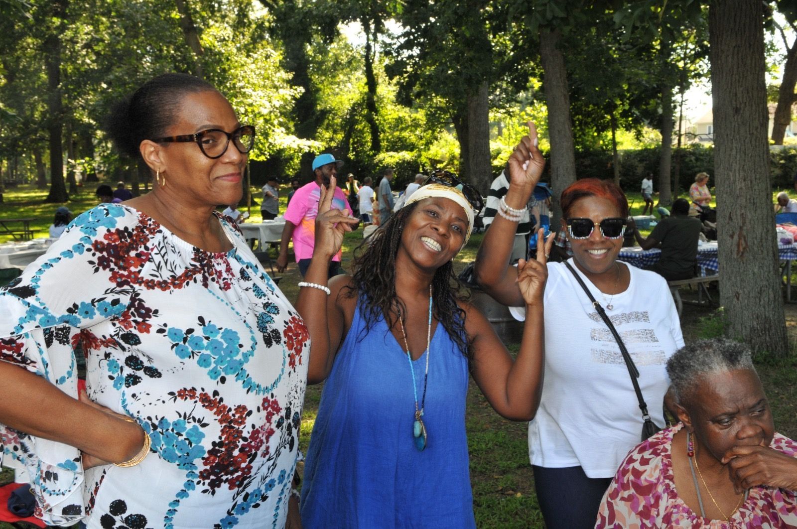 Four people smiling and posing in a park, one raising a hand, with trees and other people in the background