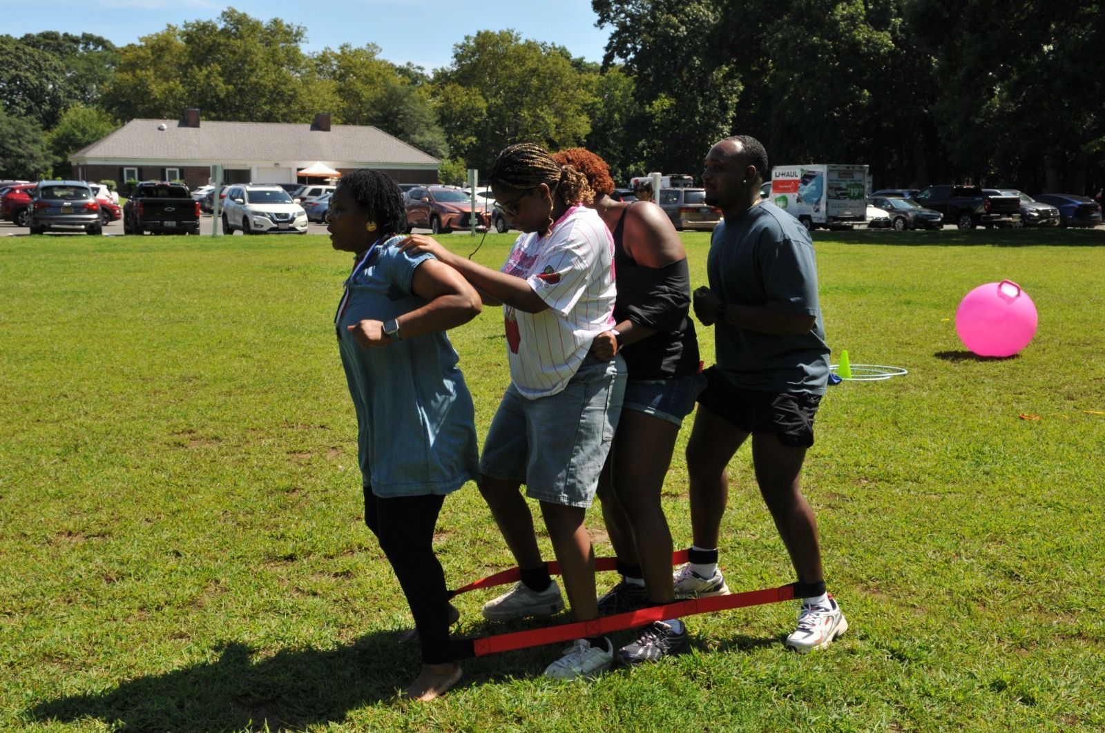 Three people balancing on a red board during an outdoor team activity on a grassy field.