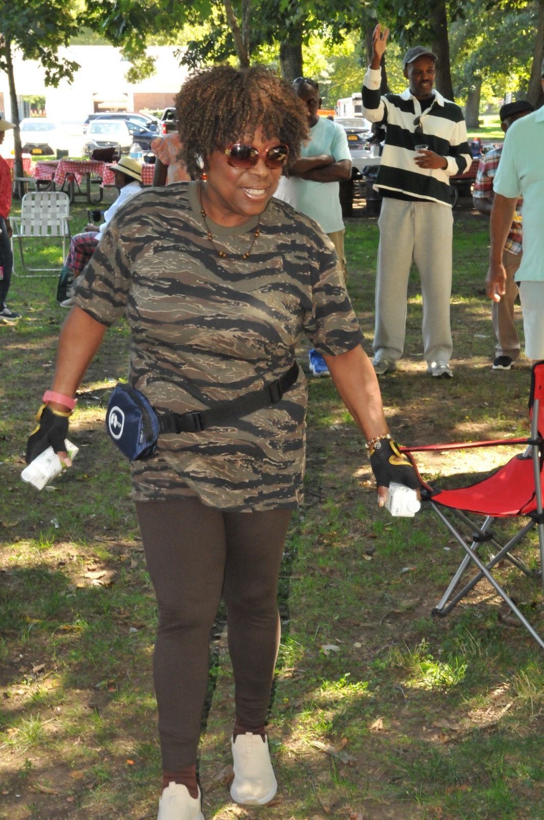 Woman walking outdoors in a park, wearing sunglasses and camouflage top, with folding chairs nearby.