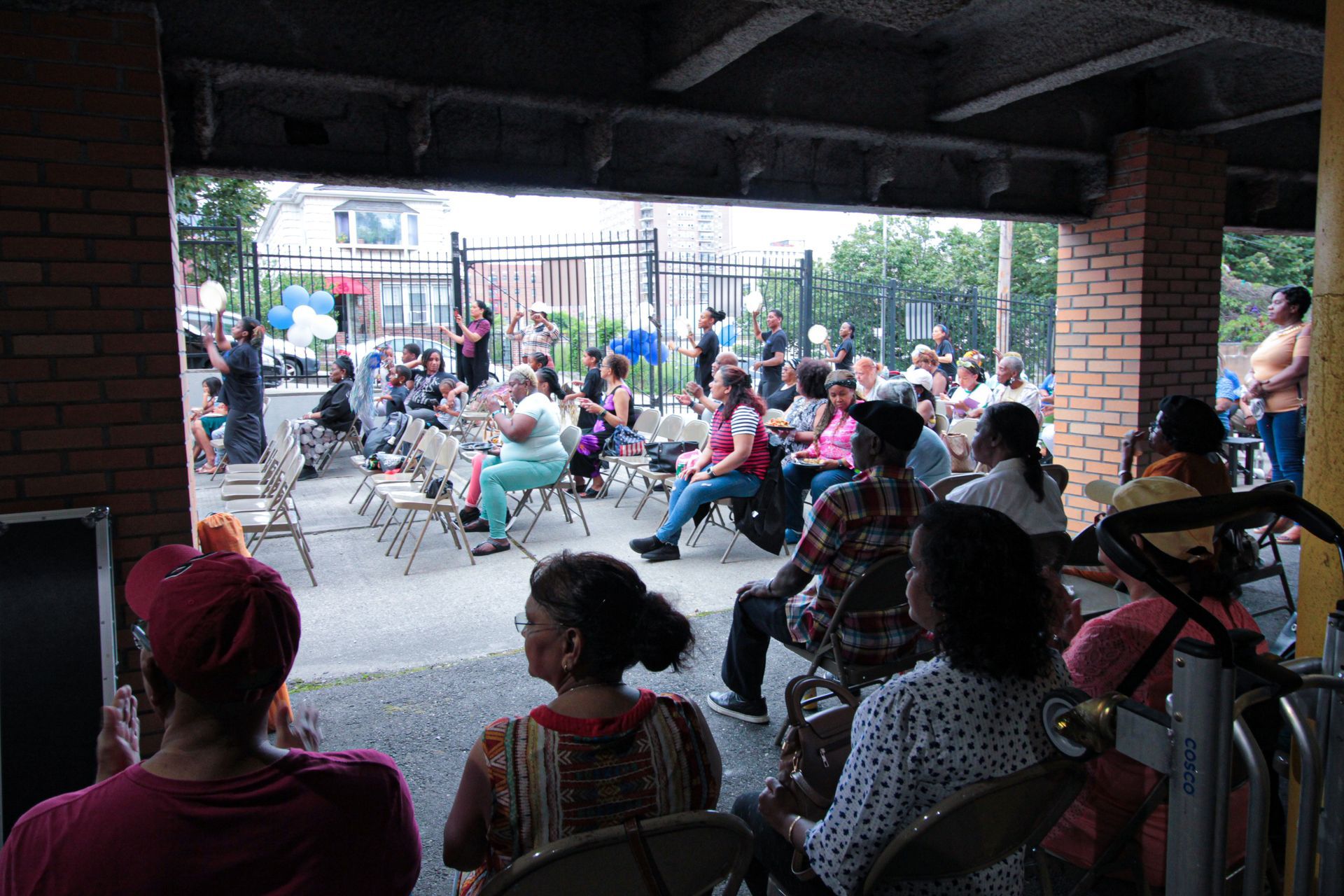 A group of people are sitting in folding chairs under a brick building.