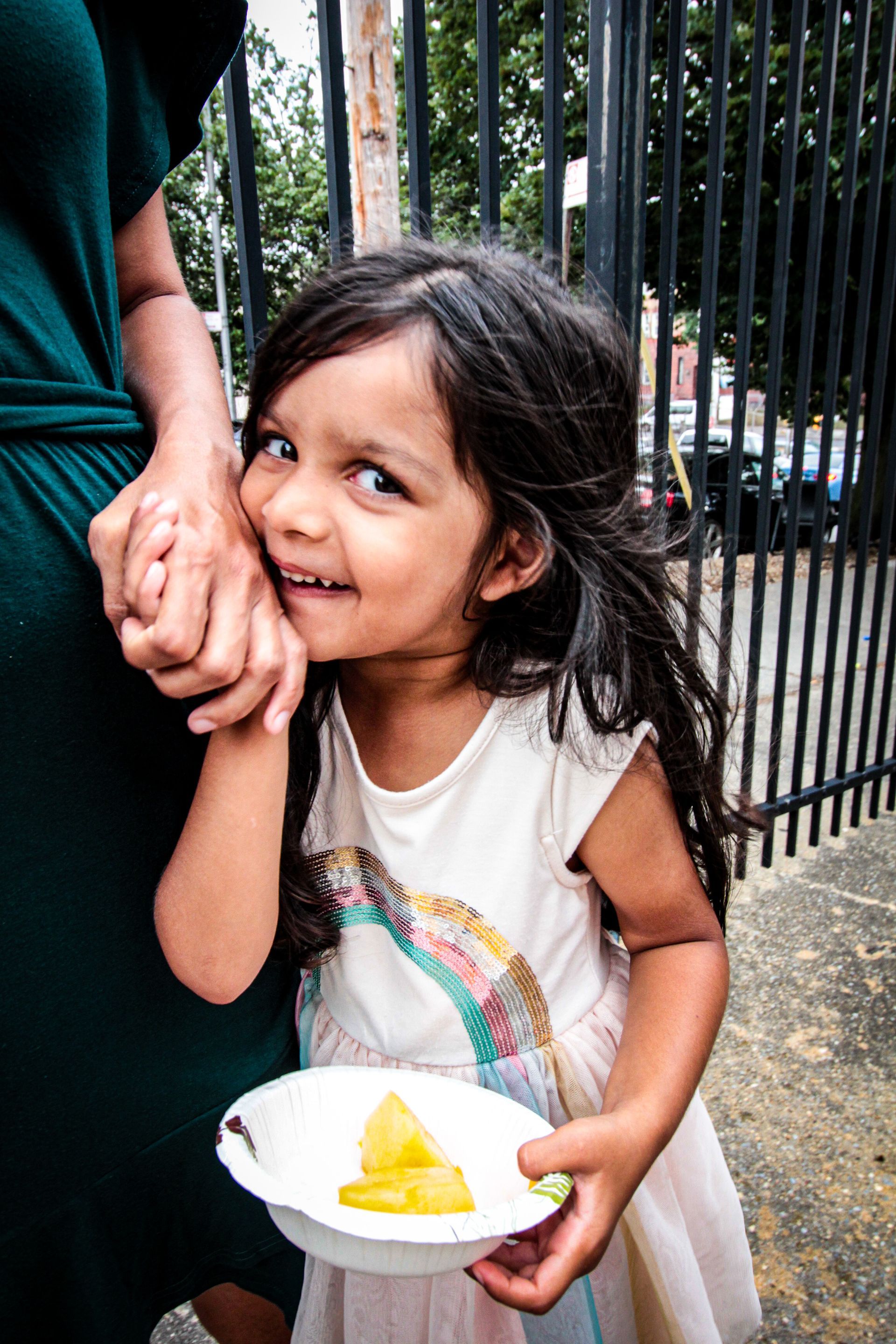 A little girl is holding a bowl of food and smiling while standing next to a woman.