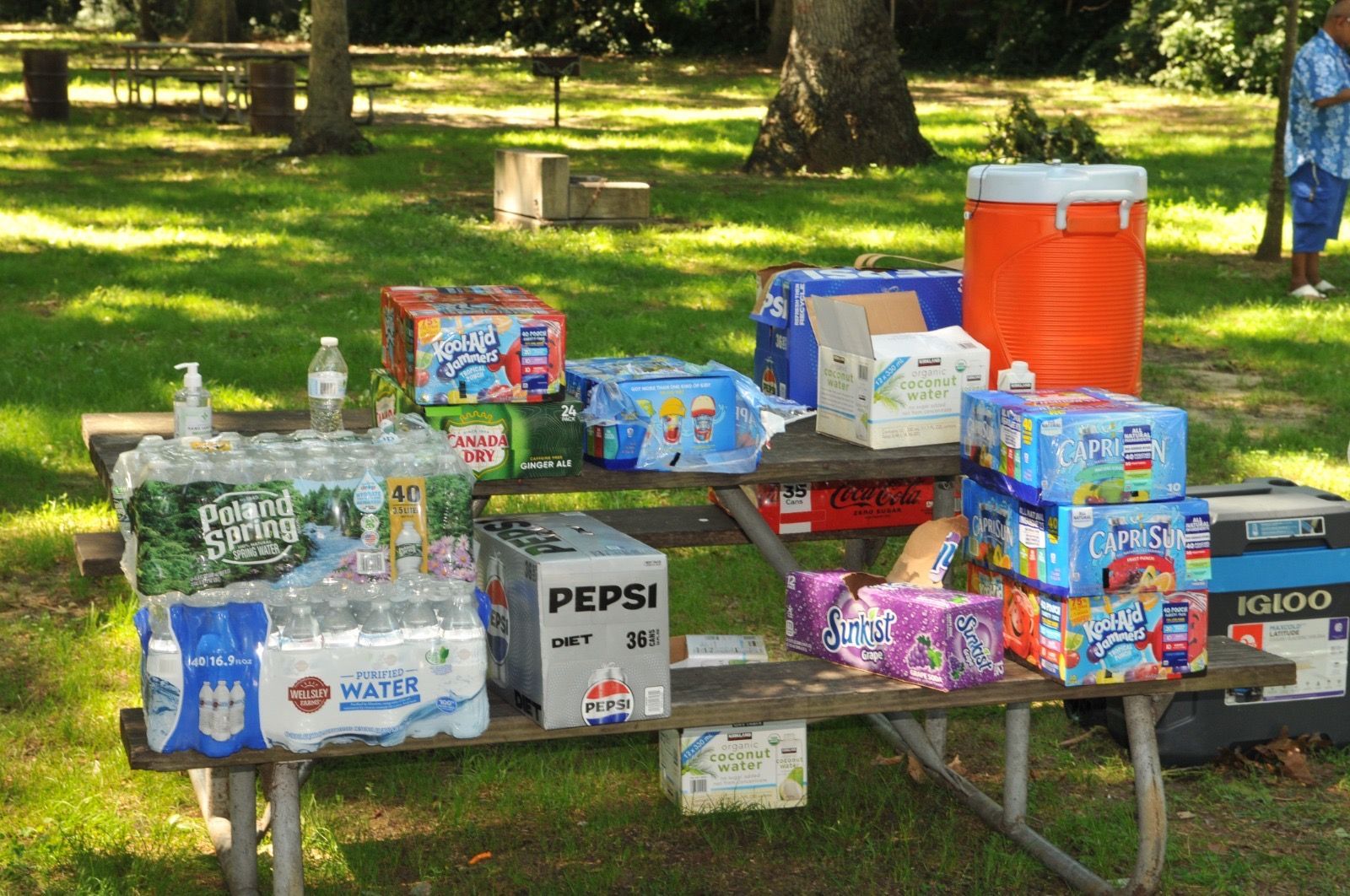 Picnic table covered with snack and drink boxes, water bottles, and a large orange cooler in a grassy park.