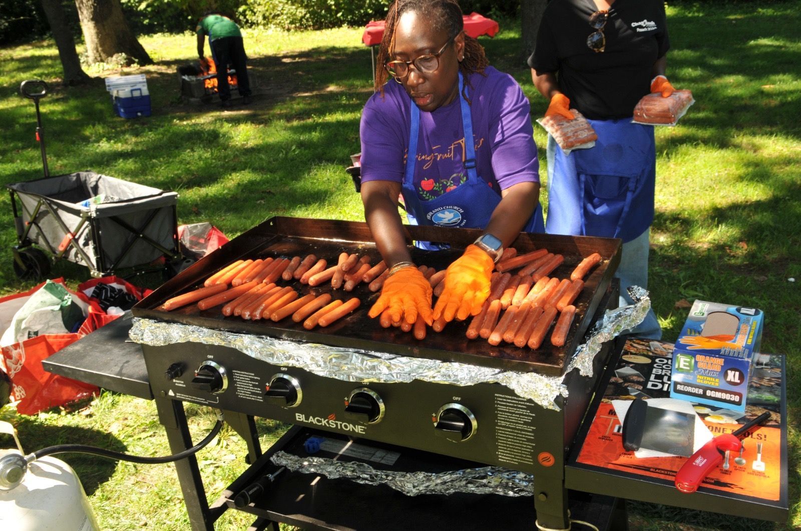 Person grilling many hot dogs on a large outdoor grill at a park picnic