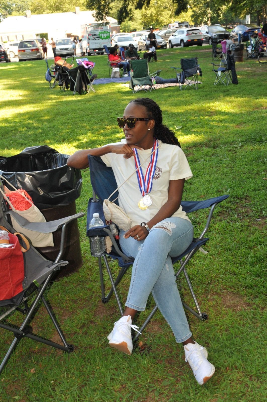 Woman seated on a folding chair in a grassy park, wearing sunglasses and medals around her neck