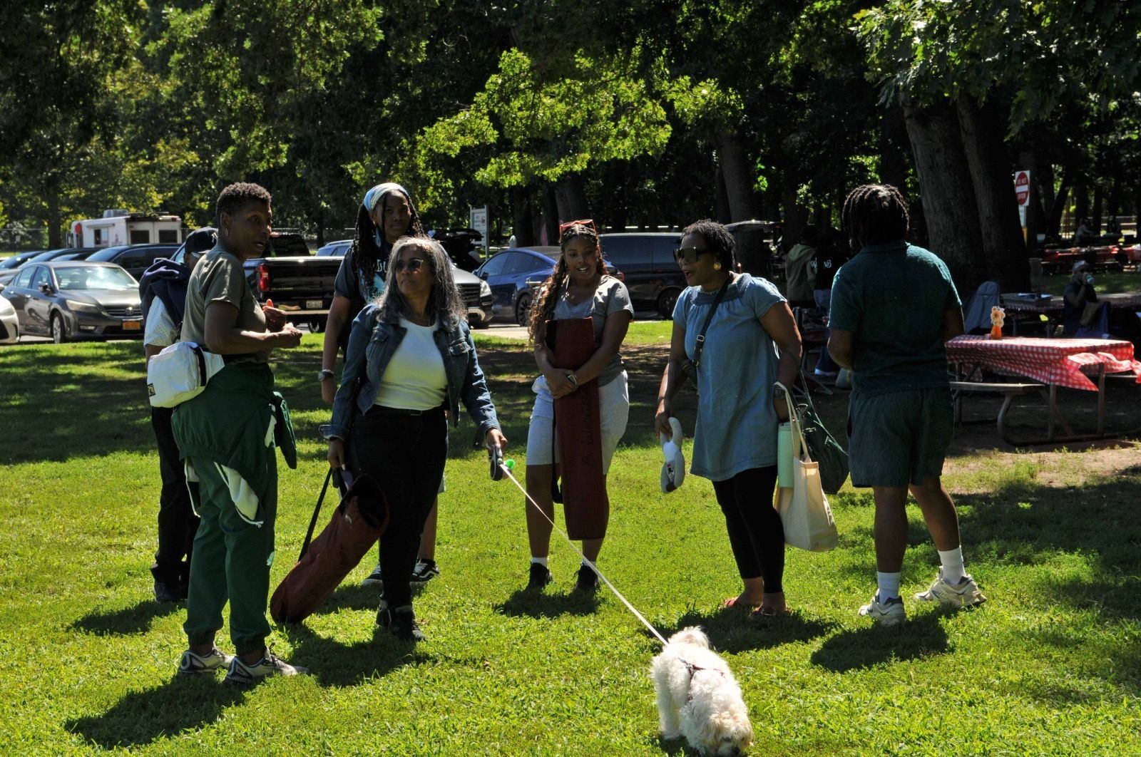 People standing on a sunny lawn with backpacks, trees and parked cars in the background; small dog in front